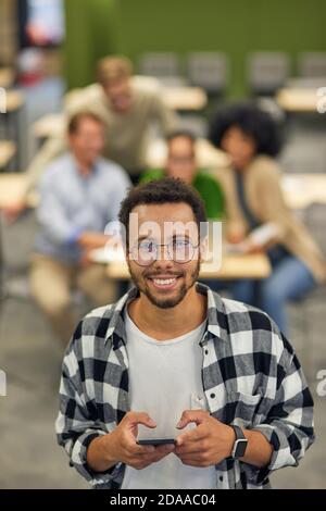 Chatten mit Freunden. Junger glücklicher Mischling hält Smartphone und lächelt vor der Kamera, während er im Coworking Space mit Kollegen im Hintergrund steht. Selektiver Fokus Stockfoto