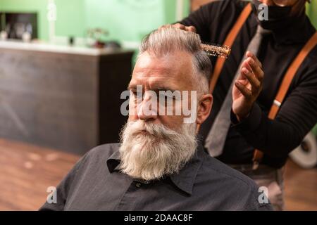 Der junge afroamerikanische Barbier und Dreadlocks mit einer Maske zur Prävention von Coronaviren kämmt mit einem Kamm und seinen Händen einen reifen weißen Kaukasier Stockfoto