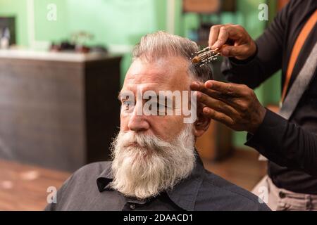 Der junge afroamerikanische Barbier und Dreadlocks mit einer Maske zur Prävention von Coronaviren kämmt mit einem Kamm und seinen Händen einen reifen weißen Kaukasier Stockfoto