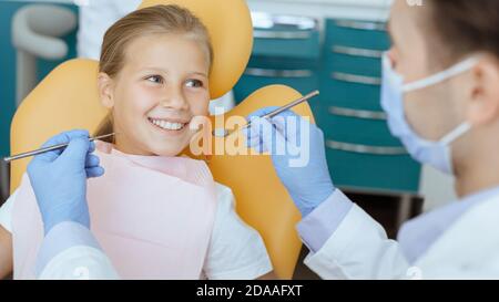 Arzt in Schutzmaske, weißem Mantel und Gummihandschuhe mit Werkzeugen untersucht kleine lächelnde Kind im Stuhl Stockfoto