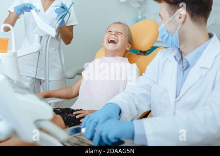 Junge Ärztin und Krankenschwester in Schutzmasken, Handschuhen und Uniformen schaut auf lachendes Mädchen Stockfoto
