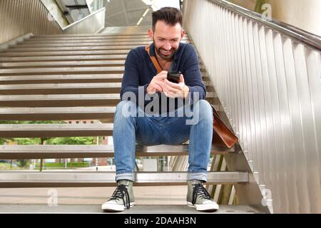 Portrait von lächelndem Mann mittleren Alters, der draußen auf Stufen sitzt Und SMS auf dem Mobiltelefon lesen Stockfoto