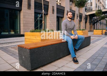Breite Aufnahme des jungen bärtigen gutaussehenden Mannes, der auf der städtischen Bank an der modernen europäischen Stadtstraße sitzt. Stockfoto