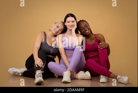 Multiethnische Freundinnen in Sportbekleidung posieren im Studio, sitzen auf dem Boden Stockfoto