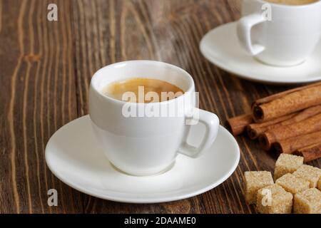 Espresso in einer weißen Tasse dampfen, Zimt und brauner Zucker auf einem Holztisch. Selektiver Fokus. Stockfoto