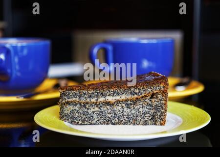 Nahaufnahme Stück Mohnkuchen auf einer Untertasse gegenüber Tassen Von Kaffee oder Tee auf einem dunklen unscharfen Hintergrund Stockfoto