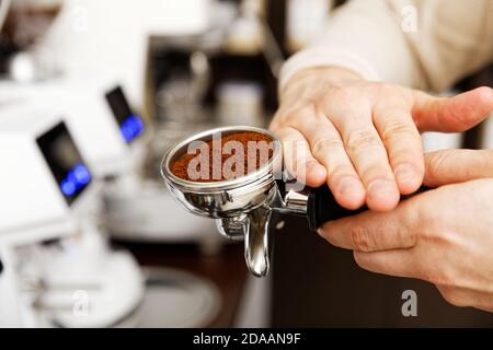 Hände des Barista halten einen mit gemahlenem Kaffee gefüllten Portafilter und bereiten sich auf die Zubereitung mit der Kaffeemaschine vor. Geringer Fokus und unscharfer Hintergrund. Stockfoto