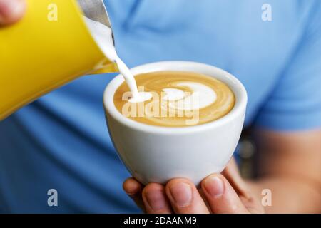 Weiße Tasse Cappuccino aus der Nähe im Café. Barista hält und gießt Milch, um Latte Art zu machen. Flacher Fokus. Stockfoto