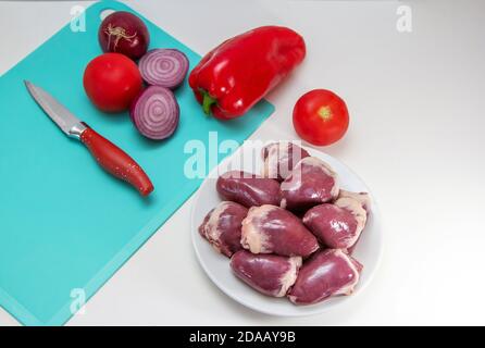 Frische rohe putenherzen auf einem Teller. Vogelgiblets bereit zum Kochen mit Gemüse – Paprika, Tomate und rote Zwiebel Stockfoto