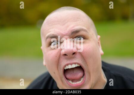 Junger Mann mit rasiertem Kopf, der seine Dominanz bedrohlich brüllt An der Kamera in Frustration im Freien in einem Park Stockfoto