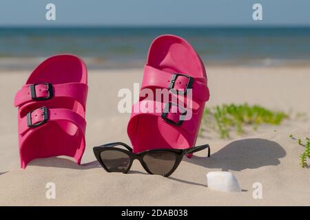 Rosa Sandalen, Sonnenbrillen und Sonnencreme am Strand an einem schönen sonnigen Tag. Pantoffeln im Sand am Meer. Flip Flops am Ufer am Meer. Stockfoto