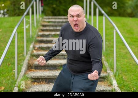 Wütender junger Mann, der die Kamera anschreit, während er steht Blockieren einer äußeren Reihe von Schritten in einem Park schwingt Seine Fäuste im Zorn Stockfoto