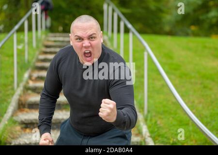 Wütender Mann, der die Kamera mit der Faust bedroht Schreit Missbrauch mit einem heftigen Ausdruck auf draußen tritt in Ein Park Stockfoto