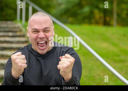 Frustrierter, wütender Mann, der wütend auf die Kamera schreit Und seine Fäuste in einer bedrohlichen Geste im Freien festklammend Ein Park Stockfoto