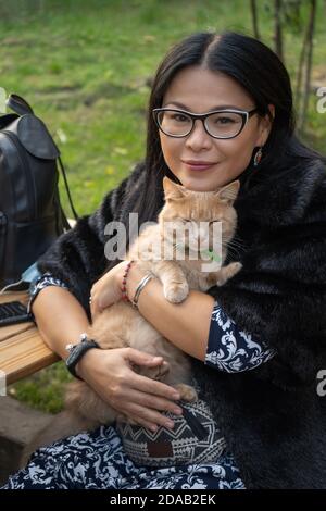 Frau mittleren Alters umarmt Katze, während sie sich auf der Straßenbank im Freien ausruhen. Glückliche reiche asiatische Frau in schwarzem Pelz Mantel mit Ingwer Katze in den Armen Stockfoto