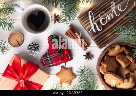 Startseite Schriftzug verbranntes Holzschild, Lebkuchenkeks, Christbaumzweige, Tasse Kaffee, Geschenk, Zimtstangen auf weißer Oberfläche. Flach liegend Stockfoto