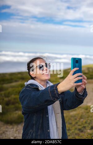 Junge Frau macht ein Selfie mit ihrem Handy auf einem Berg inmitten der Natur. Frau in Sonnenbrille lächelnd. Stockfoto