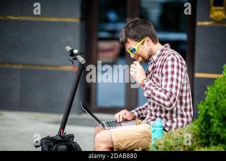 Junge kaukasischen Mann arbeiten auf Laptop, während essen nahrhafte Bar, sitzen auf der Bank in der Nähe Elektroroller im Hof. Gesund, Fastfood. Müsliriegel Stockfoto