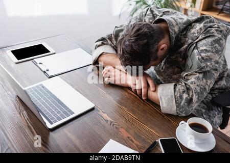 Militärmann in Uniform liegt auf dem Schreibtisch in der Nähe von Laptop und Gadgets mit leerem Bildschirm Stockfoto