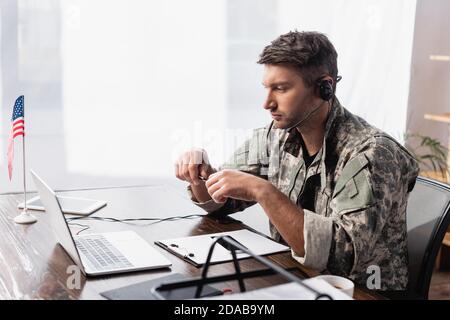 Patriotischer Militärangehörige im Headset, der Stift hält und anschaut Laptop in der Nähe der amerikanischen Flagge Stockfoto