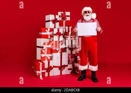 In voller Länge Foto von im Ruhestand Opa grauen Bart Stapel geschenkboxen Halten Plakette leeren Raum tragen santa Kostüm Hosenträger Sonnenschutzstiefel Gestreiftes Hemd Stockfoto