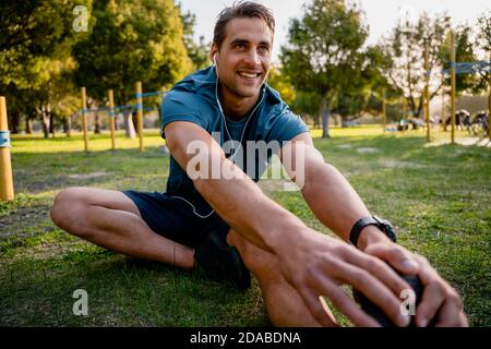 Passen Sie jungen männlichen Athleten Stretching Beinmuskulatur vor dem Laufen in Outdoor-Park hören Musik in Ohr-Handys Smart Watch Für Aktivität Stockfoto