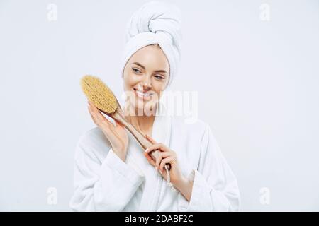 Schöne frohe Frau mit gesunder Haut und toothy Lächeln hält Holzmassagebürste für Körper, trägt Bademantel und gewickeltes weiches Handtuch auf Kopf, steht ag Stockfoto