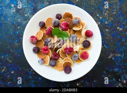 Teller mit Mini-Pfannkuchen und Beeren auf dunkelblauem Hintergrund. Flache Lagen. Stockfoto