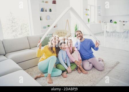 Ganzkörper-Foto von Harmonie Familie Menschen Mama Papa sitzen Boden halten Papier Karte Dach über zwei kleine Kinder in Haus drinnen Stockfoto