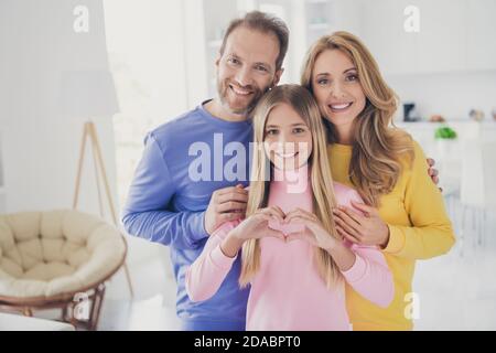 Foto von positiven fröhlichen Familie Mama Papa stehen hinter kleinen Kind Mädchen zeigen Herz Finger Zeichen im Haus drinnen Stockfoto
