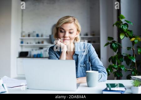 Junge blonde Frau lehnt Kinn an der Hand und arbeitet an Notebook Stockfoto