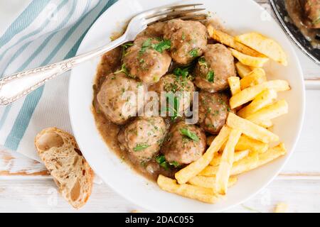 Fleischbällchen in Zwiebelsauce mit Petersilie und Pommes auf einem weißen Holztisch. Draufsicht Stockfoto