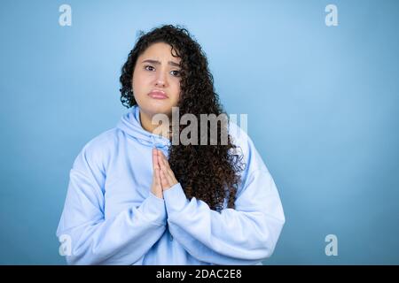 Junge schöne Frau trägt lässige Sweatshirt über isolierten blauen Hintergrund Betteln und Beten mit Händen zusammen mit Hoffnungsausdruck auf Gesicht ver Stockfoto