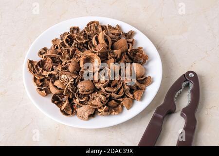 Stapel von leeren gebrochenen Walnussschalen auf einem weißen Teller und Nussknacker über Küchentisch. Biologisch abbaubare Lebensmittelabfälle und Peelings. Konzept der Nussernte. Stockfoto