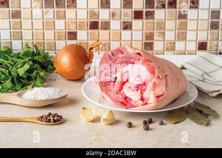 Zutaten für Aspik oder Gelied-Fleisch: Schweineschenkel auf einem Teller, Zwiebel, Knoblauch, Salz, Pfeffer, Petersilie und Lorbeerblätter auf dem Tisch. Hausgemachtes Essensrezept. Stockfoto