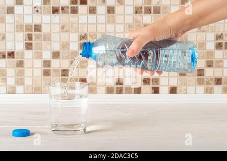 Frau Hand gießt reines Trinkwasser aus einer blauen Plastikflasche in ein Glas auf einem weißen Holztisch. Durst löschen und Gesundheitskonzept. Vorderansicht. Stockfoto