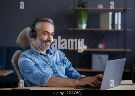 Porträt eines hübschen grauhaarigen Mannes mit Kopfhörern am Arbeitsplatz. Leitender freiberuflicher Mitarbeiter, der im Heimbüro mit einem Laptop arbeitet. Studieren Stockfoto