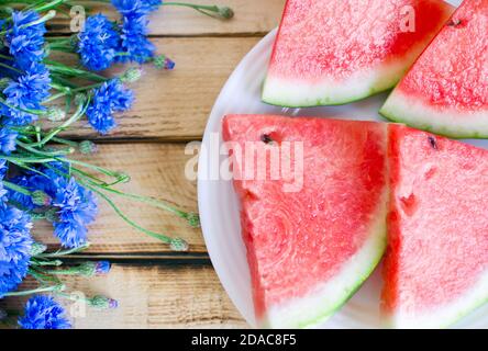 Saisonale Sommerblumen (blaue Kornblumen) und Früchte (Wassermelone und Erdbeeren) Auf rustikalem verbrannten Holztisch Stockfoto