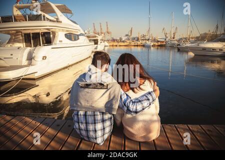 Rückansicht eines jungen Paares, das zusammen auf einem hölzernen Pier im Hafen mit kleinen Yachten in der Nähe des Meerwassers sitzt. Stockfoto