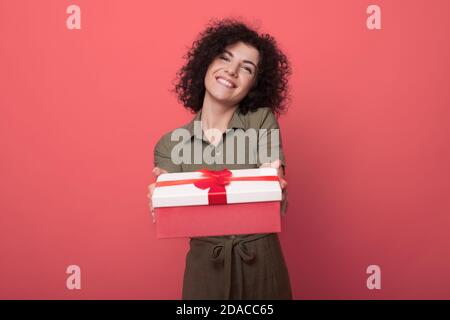 Lockig behaarte Brünette Frau trägt ein Kleid und zeigt Bei der Kamera ein Geschenk lächelnd zahnig Stockfoto