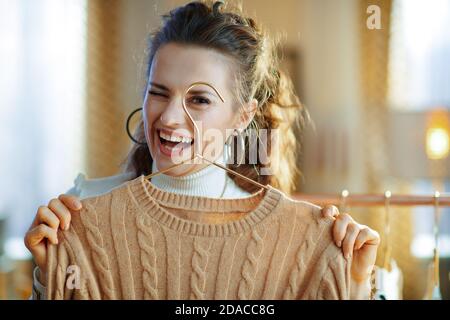 Fröhliche elegante Mittelalterhausfrau in weißem Pullover und Rock Im modernen Wohnzimmer im sonnigen Wintertag Gute Laune hält Pullover auf Kupfer Stockfoto