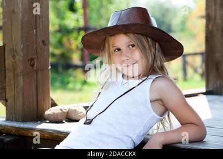 Vorschule Mädchen gekleidet ärmellose Sport-Shirt und braunen Cowboy Hut Sitzen auf Holzveranda von Holzhaus Stockfoto