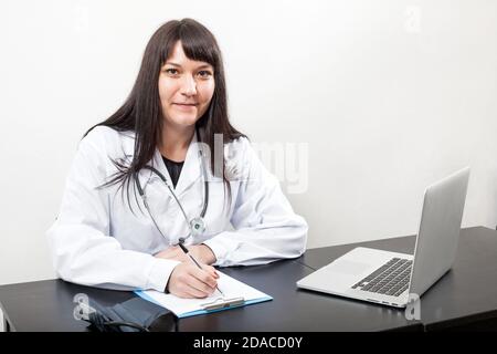 Arzt Frau bei der Arbeit sitzen am Schreibtisch im Krankenhaus oder in der Klinik, weißer Mantel, Tisch mit Laptop und Zwischenablage, Blick auf die Kamera Stockfoto