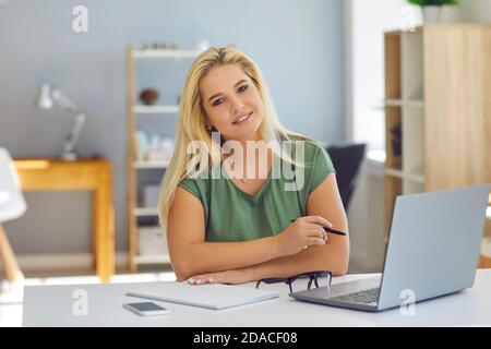 Lächelnde Frau, die online mit dem Notebook arbeitet und die Kamera anschaut Zu Hause oder im modernen Büro Stockfoto