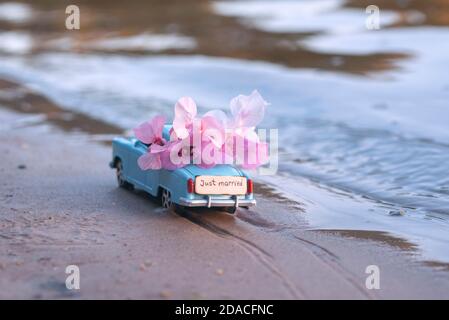 Winziges Spielzeug Retro Auto voller Blumen und Holzplatte Mit dem Text 'Just married' statt Nummernschild fährt Am Sandstrand des blauen Meeres in Stockfoto