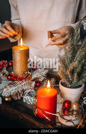 Frau Hände Blitz große goldene Kerze mit Streichhölzern in Weihnachtskranz. Weihnachten Innendekoration auf rustikalem Holzhintergrund. Hygge, Ornamente, Kerze Stockfoto