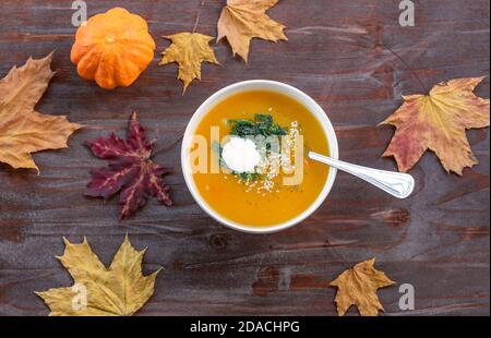 Draufsicht auf weiße Schüssel mit hausgemachter Kürbiscremesuppe, gewürzt mit Joghurt, gefrorener Petersilie und Sesam auf rustikalem Holztisch Stockfoto