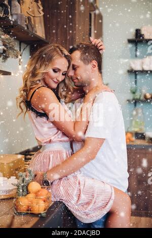 Ein Paar umarmte sich auf der Küchenunterfläche. Hübsche Blondine in rosa umarmt mit ihrem Mann oder Freund auf der Küchentheke unter Weihnachtsschnee. Stockfoto