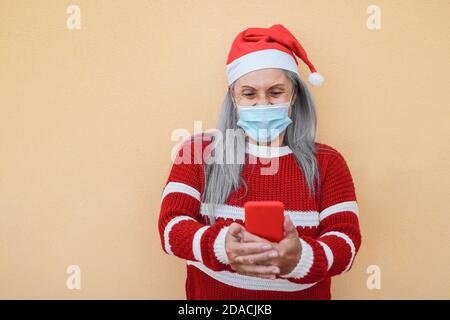 Glückliche ältere Frau trägt weihnachtsmann Hut und Gesichtsmaske Während der Verwendung von Mobiltelefonen - Fokus auf Gesicht Stockfoto