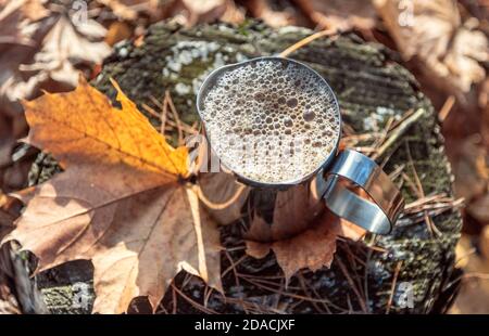 Edelstahltasse mit weißem Kaffee, bedeckt mit Milchschaum und Blasen an einem Stumpf aus nächster Nähe an einem sonnigen Herbsttag. Stockfoto
